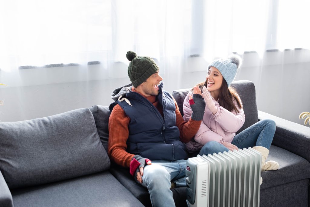 Happy couple in winter wear near a heater suggests need for Heating Contractor DC to ensure efficient warmth during cold spells in the DMV.