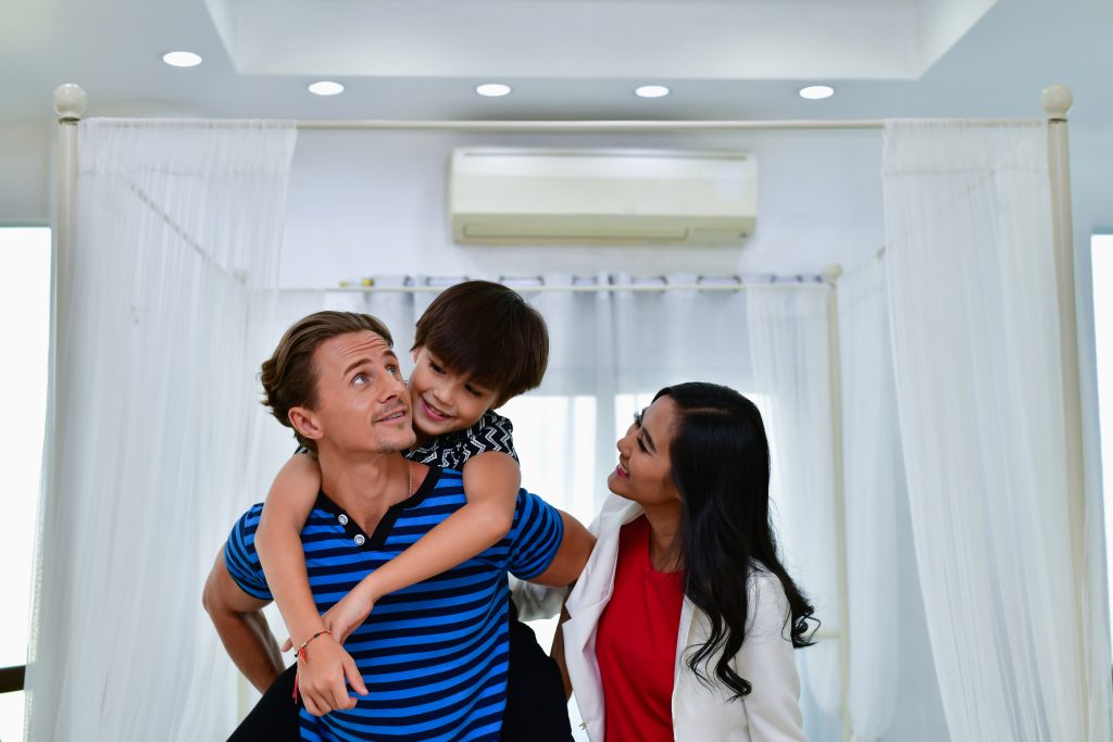 Happy family enjoying AC Repair PG comfort indoors, with a dad giving his son a piggyback ride and mom smiling at them. Cool home environment.