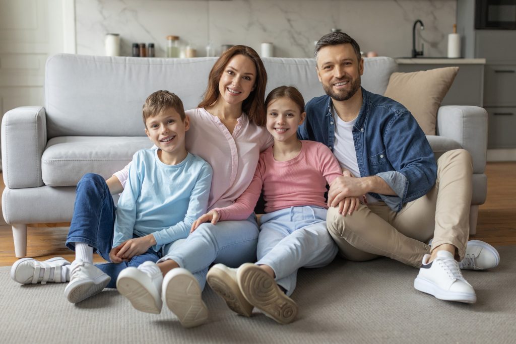 Happy family of four sitting together in their living room, smiling, near a sofa. Consider a programmable thermostat in your DMV home.